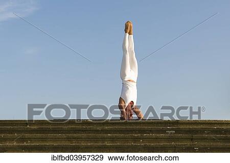 Stock Photo - "Young woman practising Hatha yoga, outdoors, showing the pose Sirshasana, Head stand". Fotosearch