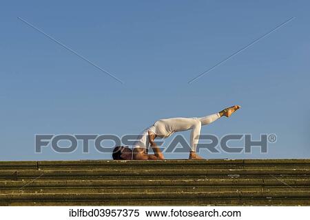 Stock Photography - "Young woman practising Hatha yoga, outdoors, showing the pose variation of Halasana, Supta konasana, variation of Plough". Fotosearch