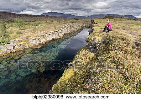 Stock Photo - "Young woman sitting next to a river, Pingvellir, Pingvellir, Iceland, Europe". Fotosearch