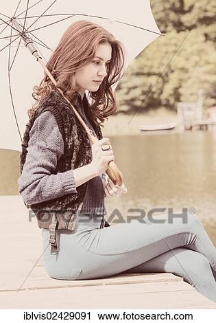 Stock Image - Young woman sitting on a jetty holding a white parasol. Fotosearch
