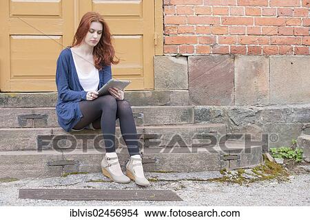 Picture - Young woman sitting on stairs in front of building holding a tablet pc. Fotosearch