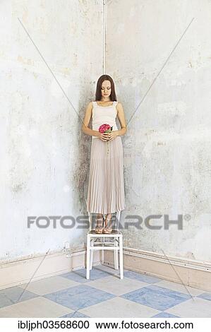 Stock Image - "Young woman standing on a chair against a wall with peeling paint, Germany, Europe". Fotosearch