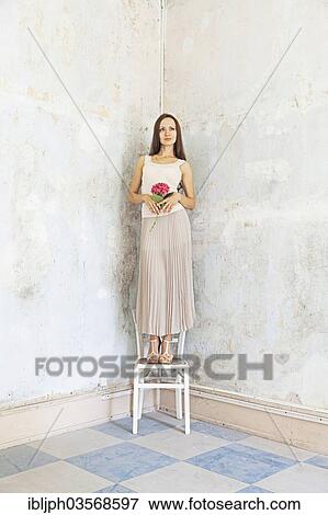 Stock Photo - "Young woman standing on a chair against a wall with peeling paint, Germany, Europe". Fotosearch