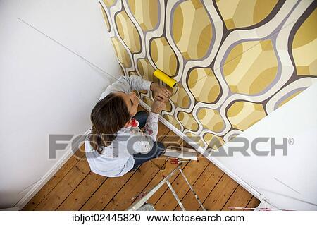 Stock Image - Young woman wallpapering a wall. Fotosearch