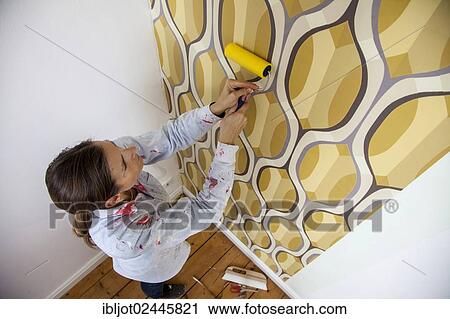 Stock Image - Young woman wallpapering a wall. Fotosearch