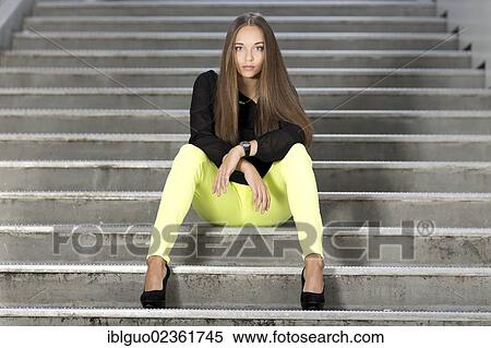 Stock Photography - "Young woman wearing a black top, bright yellow trousers and high black shoes posing while sitting on stairs". Fotosearch