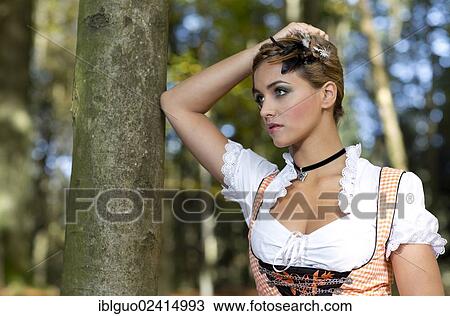 Stock Image - Young woman wearing a dirndl dress posing against a tree, portrait, dirndl look. Fotosearch