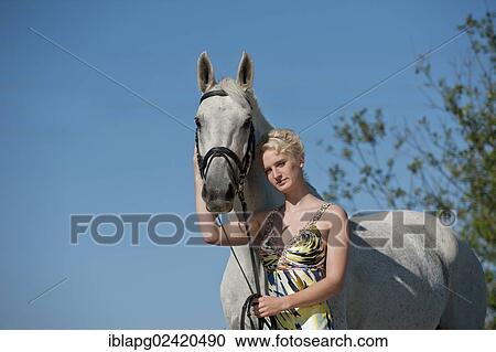 Stock Image - Young woman wearing a dress standing with a Hanoverian horse. Fotosearch