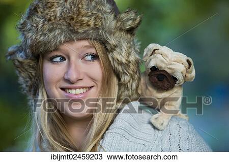 Stock Image - Young woman wearing a fur hat with a plush pug on her shoulder. Fotosearch