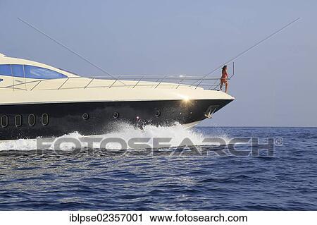 Stock Image - "Young woman wearing a red swimsuit standing on the bow of a moving motor yacht, French Riviera, Cote d'Azur, Mediterranean Sea, Europe". Fotosearch