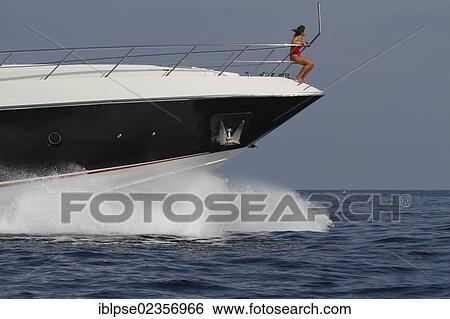 Stock Photograph - "Young woman wearing a red swimsuit sitting on the railing of the bow of a moving motor yacht, French Riviera, Cote d'Azur, Mediterranean Sea, Europe". Fotosearch