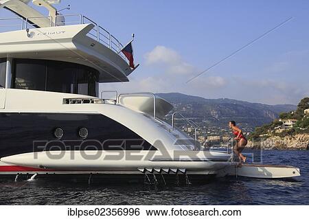 Stock Photograph - "Young woman wearing a red swimsuit climbing up a ladder from the swim platform of a motor yacht, French Riviera, Cote d'Azur, Mediterranean Sea, Europe". Fotosearch