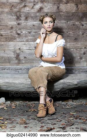 Young woman wearing a white dirndl top and traditional leather pants sitting on a wooden bench, dirndl look View Large Photo Image Stock Image - Young woman wearing a white dirndl top and traditional leather pants sitting on a wooden bench, dirndl look. Fotosearch