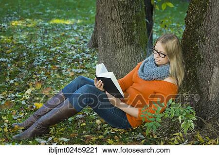 Stock Image - Young woman wearing glasses sitting in the park and reading a book. Fotosearch