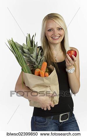 Stock Image - "Young woman with a bag full of vegetables and fruit in her arm, holding an apple in her hand". Fotosearch