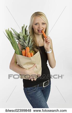 Stock Photo - "Young woman with a bag full of vegetables and fruit in her arm, holding a carrot in her hand". Fotosearch