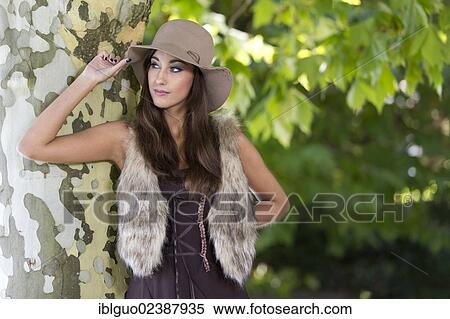 "Young woman with a hat, fake fur waistcoat and a brown dress posing at a tree" View Large Photo Image Stock Photography - "Young woman with a hat, fake fur waistcoat and a brown dress posing at a tree". Fotosearch