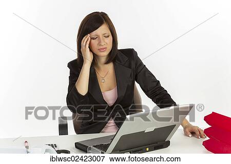 Stock Photo - Young woman with a headache sitting at a desk with a laptop. Fotosearch