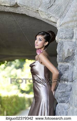 Stock Image - Young woman with an updo hairstyle and a silvery evening dress posing in front of an archway. Fotosearch