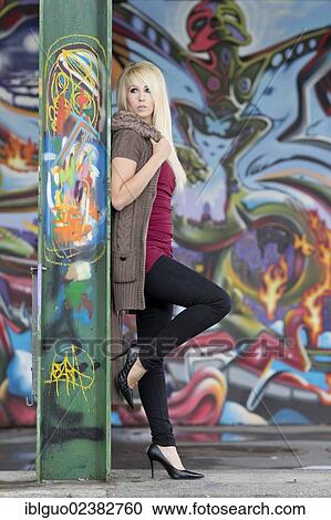 Young woman with long blond hair wearing a hooded jacket posing while leaning against a steel beam with graffiti in front of a wall with graffiti View Large Photo Image Stock Image - Young woman with long blond hair wearing a hooded jacket posing while leaning against a steel beam with graffiti in front of a wall with graffiti. Fotosearch