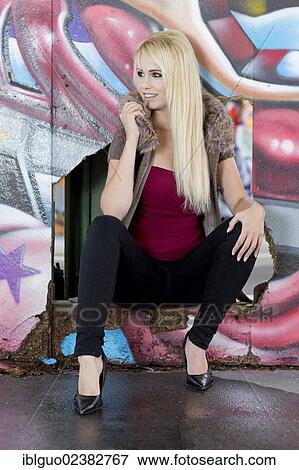 Young woman with long blond hair wearing a hooded jacket posing while crouching in front of a wall with graffiti View Large Photo Image Stock Photo - Young woman with long blond hair wearing a hooded jacket posing while crouching in front of a wall with graffiti. Fotosearch