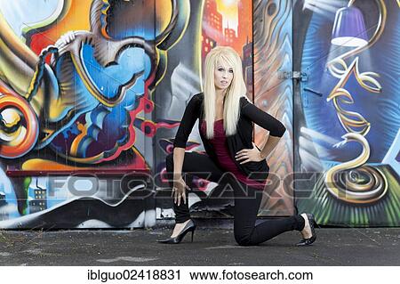 Stock Image - Young woman with long blonde hair posing in front of a wall with graffiti. Fotosearch