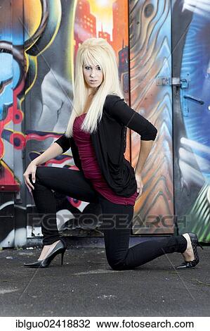 Stock Image - Young woman with long blonde hair posing in front of a wall with graffiti. Fotosearch