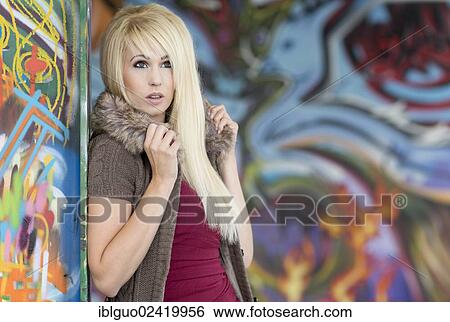 Stock Photograph - Young woman with long blonde hair posing against a steel girder in front of a wall with graffiti. Fotosearch