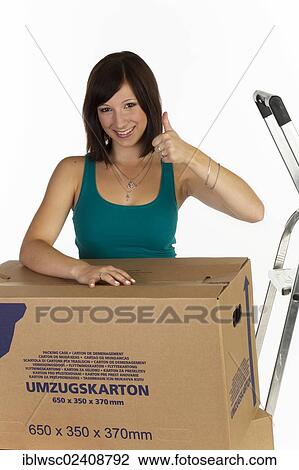 Stock Image - "Young woman with moving boxes, making a thumbs-up gesture". Fotosearch