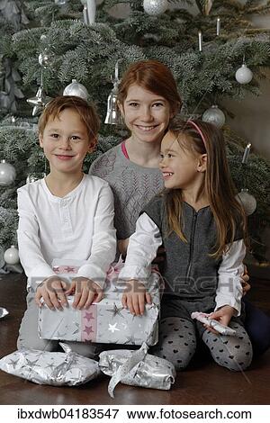Stock Photo - Christmas Eve, happy siblings with Christmas presents under the Christmas tree, Bavaria, Germany, Europe. Fotosearch