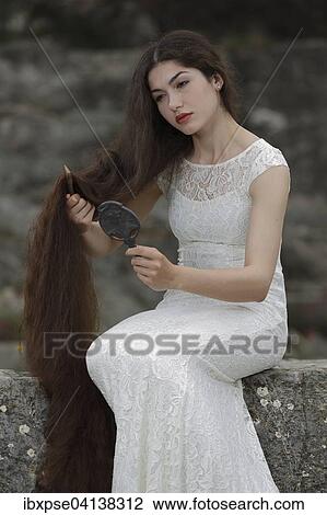 Stock Image - Enactment of the painting Lady Lilith by Dante Gabriel Rossetti, young woman in a white dress, combing her hair and looking in the mirror. Fotosearch