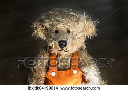 Picture - Light brown teddy bear, with orange overalls, portrait against a dark background. Fotosearch