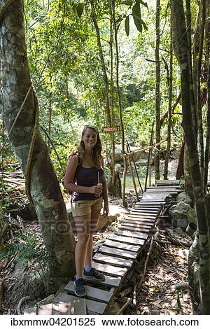 Stock Photography - Tourist smiling, holding a drink, standing on a boardwalk in the woods, path to the Na Muang waterfalls, Ko Samui, Thailand, Asia. Fotosearch
