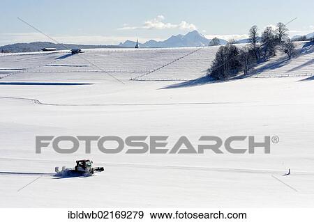 Stock Photo - Cross country ski-run track-setter and the tower of the parish church Maria Heimsuchung, Visitation of Mary, Grosshartpenning, Wendelstein mountain range at back, Holzkirchen, Upper Bavaria, Bavaria, Germany, Europe. Fotosearch