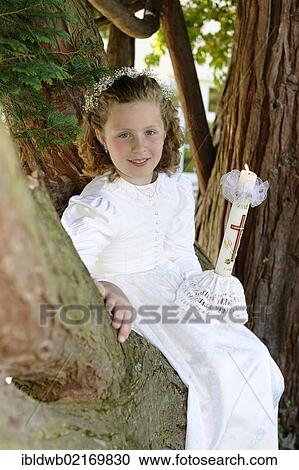 Stock Image - First communion girl with a white dress sitting on a tree. Fotosearch