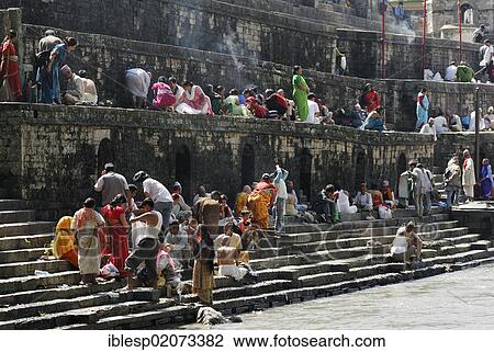 Stock Image - Pilgrims on the steps of the Pandra Shivalaya Complex opposite the Ghats of Pashupatinath on the holy Bagmati River, Kathmandu, Nepal, Asia. Fotosearch