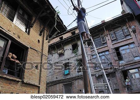 Stock Image - Young man standing on ladder that is leaning against power pole, Bhaktapur, Kathmandu Valley, Nepal, Asia. Fotosearch