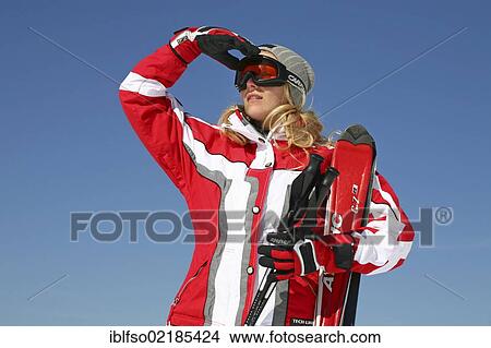 Picture - Young woman, about 25 years, wearing a red and white ski suit and a light grey cap, against a blue sky, one hand next to her ski glasses, Thuringian Forest mountains, Thuringia, Germany, Europe. Fotosearch