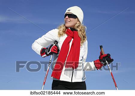 Picture - Young woman, about 25 years, wearing a bright jacket, a cap, dark sunglasses and a red scarf, against a blue sky, holding ski sticks, smiling, Thuringian Forest mountains, Thuringia, Germany, Europe. Fotosearch