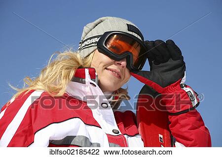 Stock Image - Young woman, about 25 years, wearing a red and white ski suit and a light grey cap, against a blue sky, one hand next to her ski glasses, Thuringian Forest mountains, Thuringia, Germany, Europe. Fotosearch