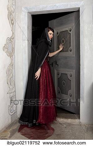 Stock Image - Young woman wearing a red dress and a black cape opening a door. Fotosearch