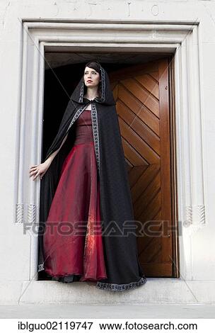 Stock Photo - Young woman wearing a red dress and a black cape standing at a door. Fotosearch