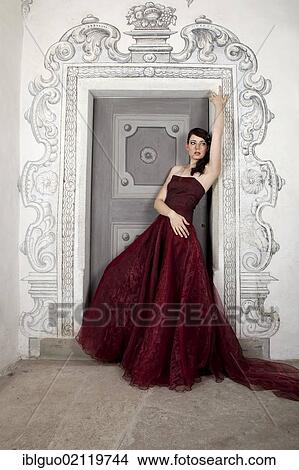 Picture - Young woman wearing a red dress with a long train leaning on a door. Fotosearch