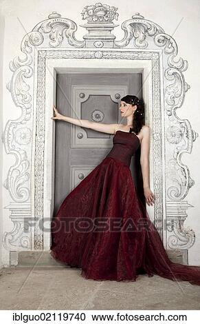 Stock Image - Young woman wearing a red dress with a long train leaning on a door. Fotosearch