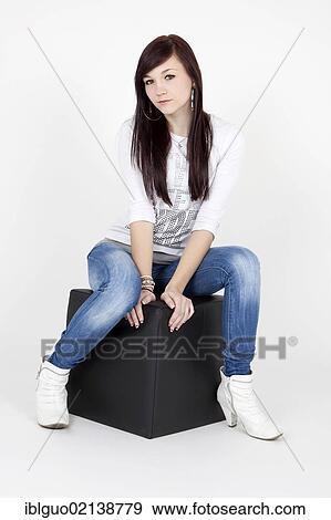 Young woman wearing a white top and jeans, posing on a black cube seat View Large Photo Image Stock Photo - Young woman wearing a white top and jeans, posing on a black cube seat. Fotosearch