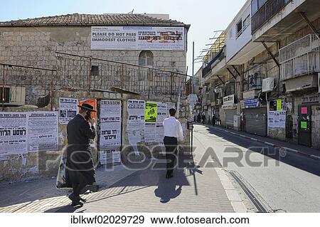 Stock Photo - Banner up on a house with rules of conduct for tourists, wall newspapers and an Orthodox Jew with a mobile phone at the front in the district Me'a She'arim or Mea Shearim, Jerusalem, Israel, Middle East, Asia. Fotosearch