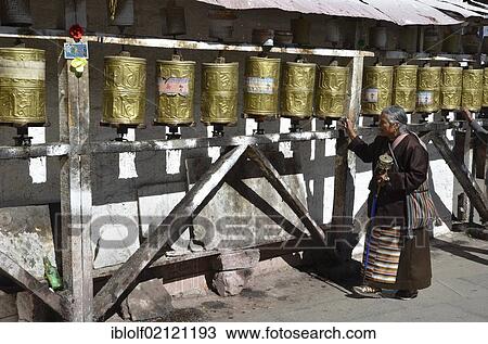 Stock Image - Elderly Tibetan woman in traditional costume surrounding a small temple, Meru Ningba, prayer wheels, Bakhor, near the Jokhang temple, Lhasa, Tibet, China, Asia. Fotosearch