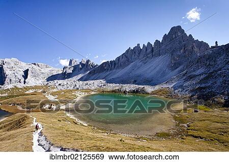 Stock Photo - Hiker descending from the Three Peaks Lodge, currently at the Boedenseen Lakes, with Paternkofel Mountain on the right, Zwoelferkofel Mountain at the rear, Alta Pusteria, Sesto, Dolomites, Alto Adige, Italy, Europe. Fotosearch