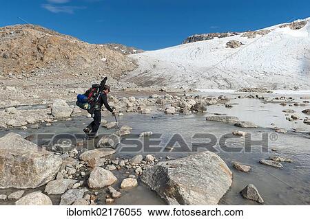 Stock Photography - Leader of a group of hikers with a backpack and a rifle at the river-crossing at Mittivakkat Glacier, Peninsula Ammassalik, East Greenland, Greenland, North America. Fotosearch