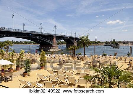 Stock Image - Leisure area with a sandy beach with deck chairs on the bank of the Rhine River, Adenauer-Ufer and Theodor Heuss Bridge, Mainz, Rhineland-Palatinate, Germany, Europe. Fotosearch